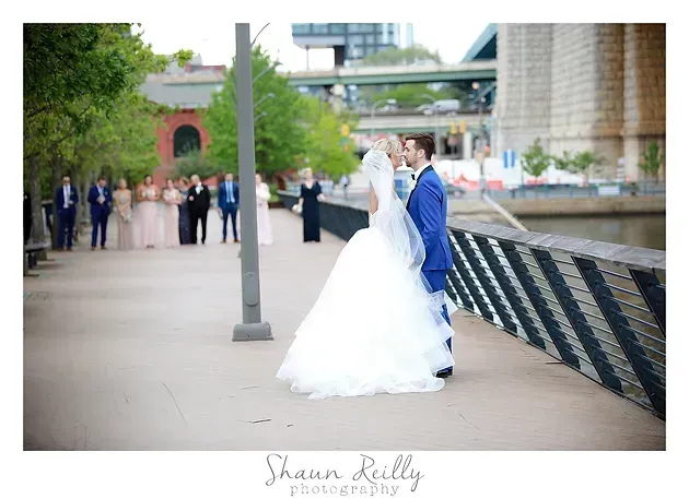 Bride and groom posing by a riverside walkway, with wedding guests in the background