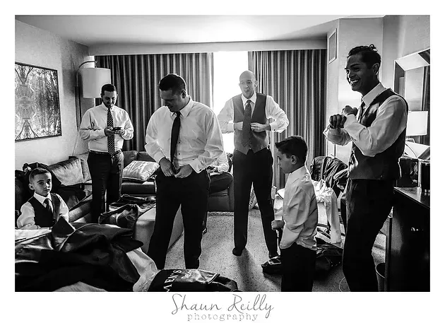 Men and boys in dress shirts and ties gather in a hotel room, some adjusting ties and holding drinks.