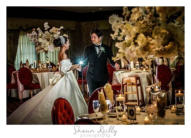 Bride and groom in a decorated reception room, lit by a camera flash, with floral arrangements and red chairs.