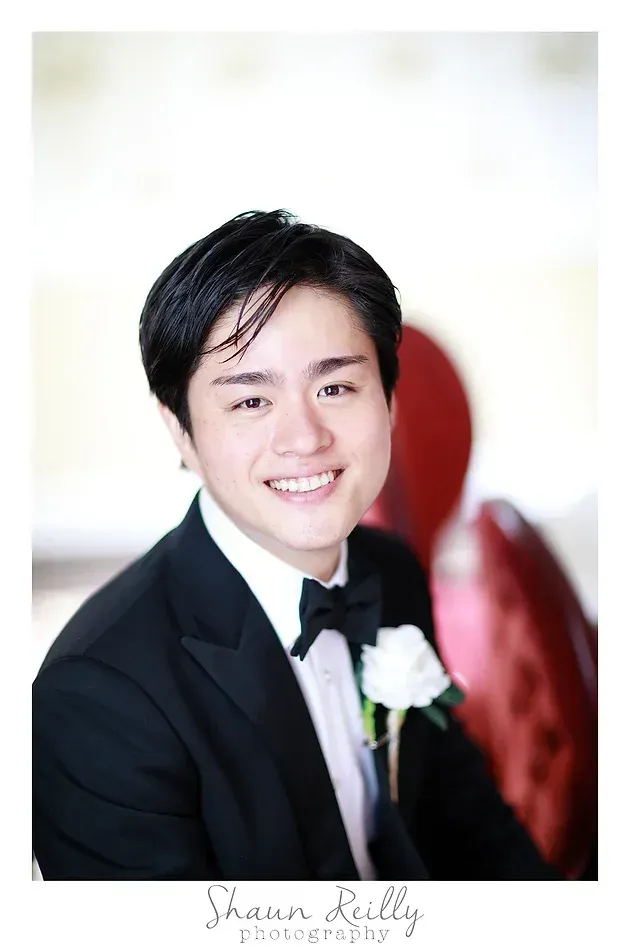 Smiling man in a black tuxedo with a white boutonniere against a light background