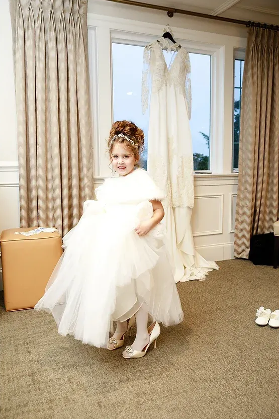 Child in a white formal dress smiling in a room, with a hanging wedding gown and curtains nearby