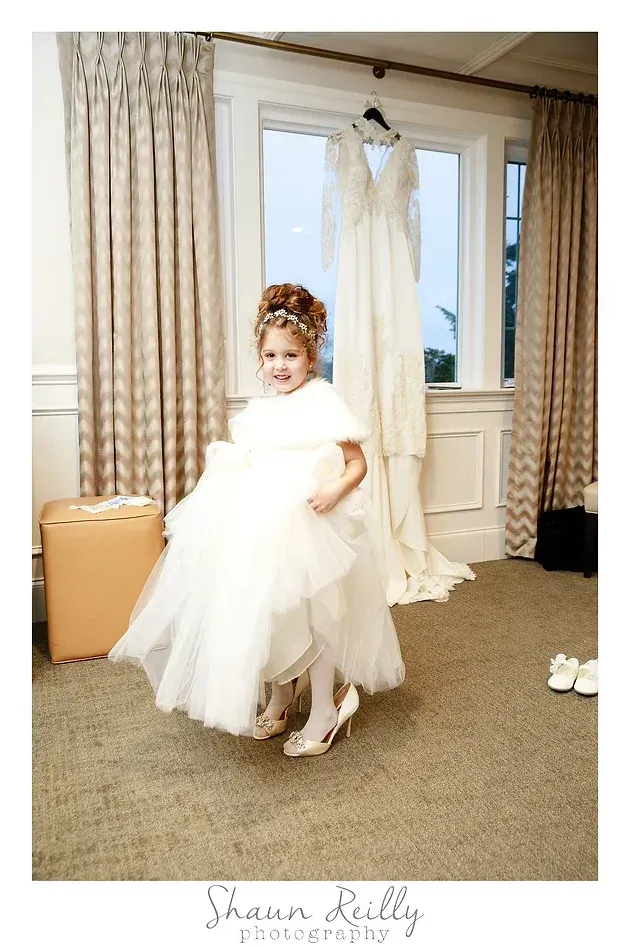 Child in a white dress and floral headpiece standing in a bridal shop near a wedding gown display.