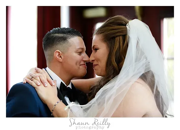 Couple in wedding attire embracing and touching foreheads, smiling in a soft-lit indoor setting