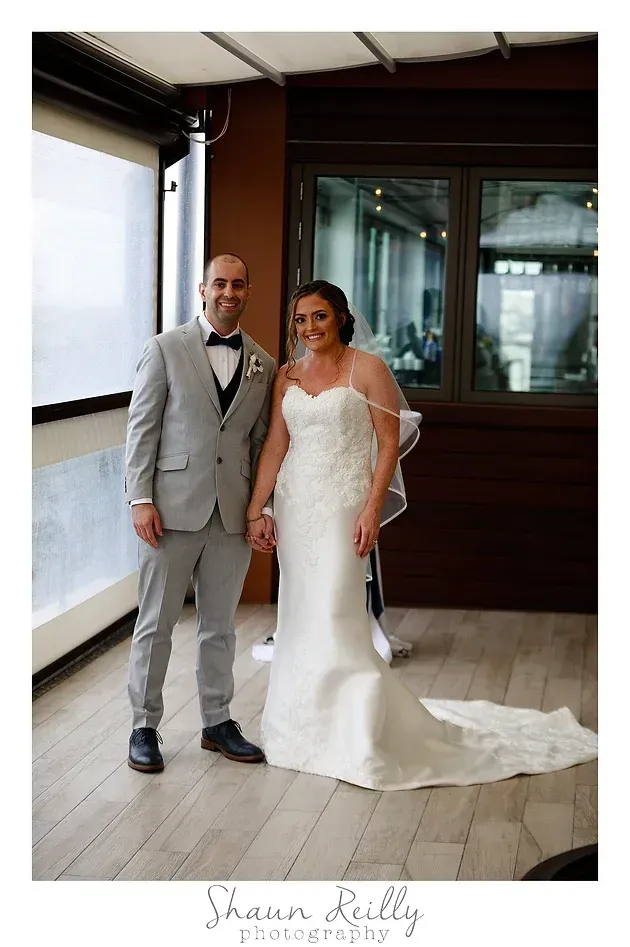 Bride and groom posing indoors, with the bride in a white wedding gown and the groom in a light gray suit.