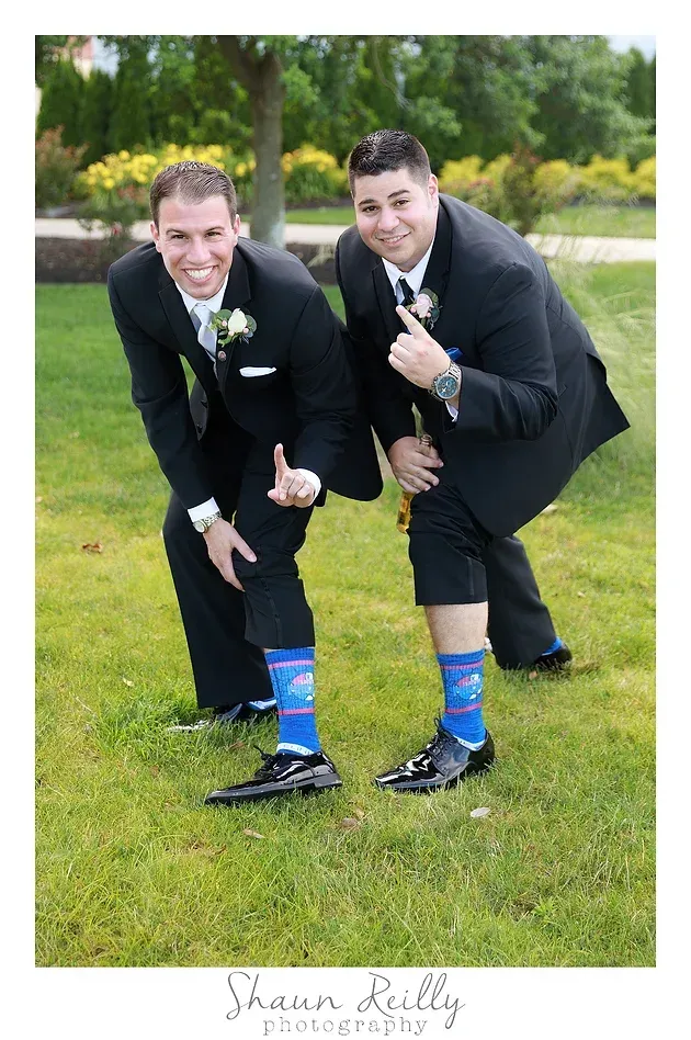 Two men in black suits crouch on a grassy lawn, wearing blue patterned socks and smiling at the camera.