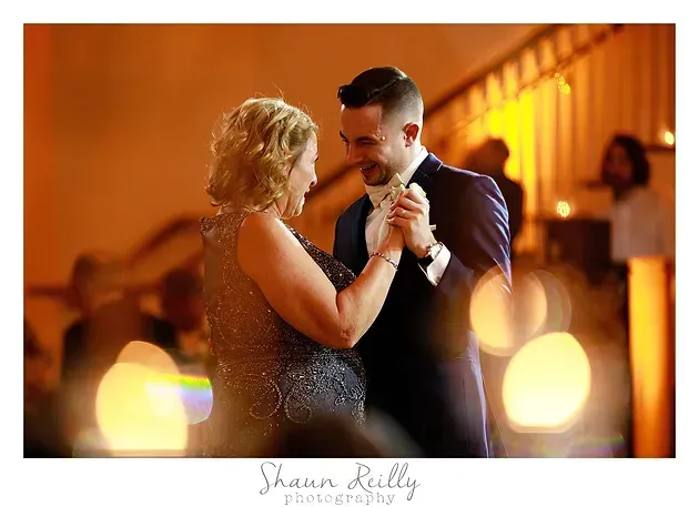 A couple dancing closely at a warmly lit reception, holding hands and smiling.