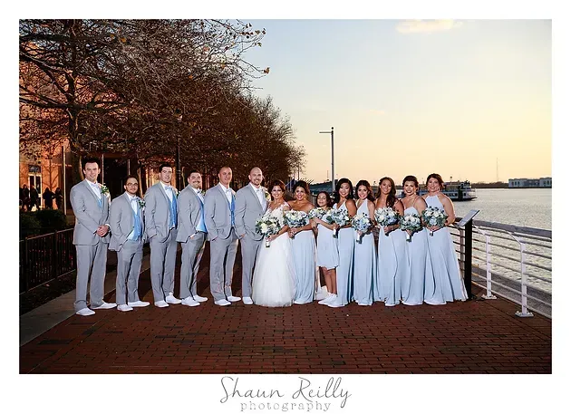 Wedding party posing by the waterfront at sunset, with bridesmaids in white dresses and groomsmen in light gray suits