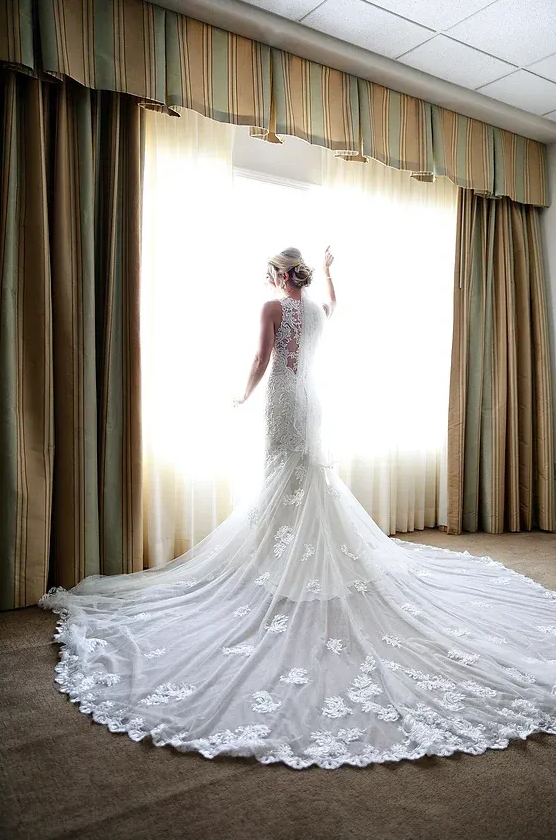 Bride in a white lace wedding gown standing before bright curtains, train spread across the floor.