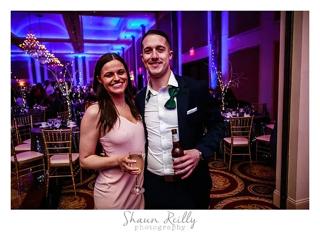 Couple posing with drinks at a purple-lit formal event in a decorated ballroom