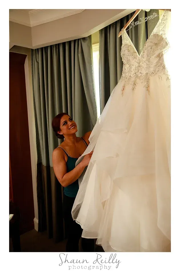 Bride adjusting a white wedding dress hanging by a window with green curtains