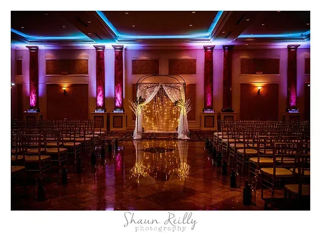 Elegant banquet hall with rows of chairs, purple uplighting, and a gold draped stage at the front.