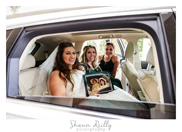 Three women smiling in a white car, holding a photo frame and looking toward the camera