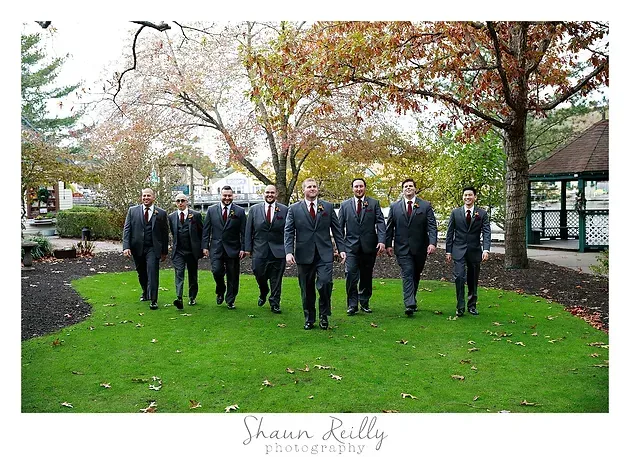 Group of people in dark suits walking on a green lawn in a park with autumn trees behind them