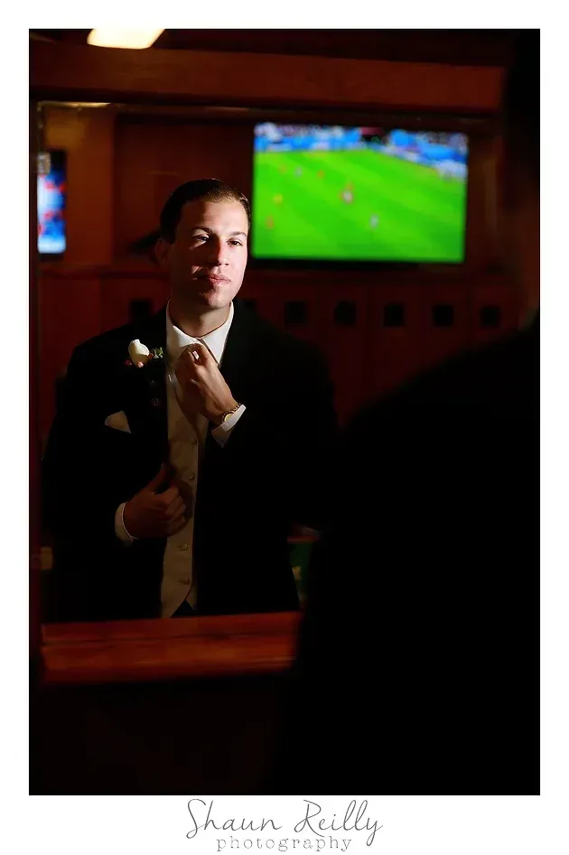 Man in a dark suit holding a microphone in a dim bar, with a TV showing a soccer match in the background