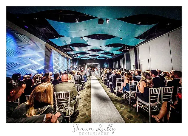 Audience seated in a modern event hall with blue geometric ceiling lights and a central aisle