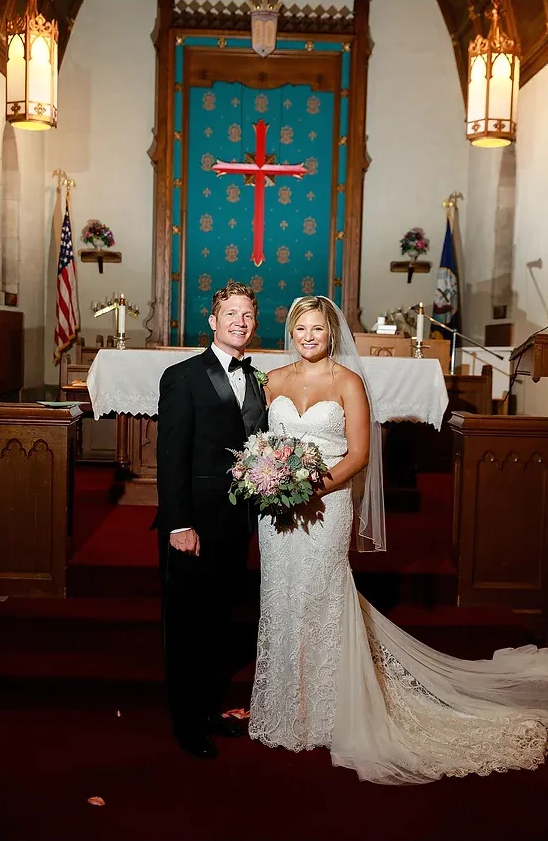 Bride and groom standing at altar in a church, holding bouquet, with red carpet and ornate decor