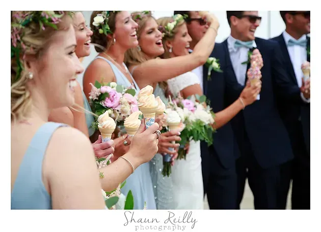 Bridesmaids and groomsmen hold bouquets at an outdoor wedding ceremony, smiling and clapping.