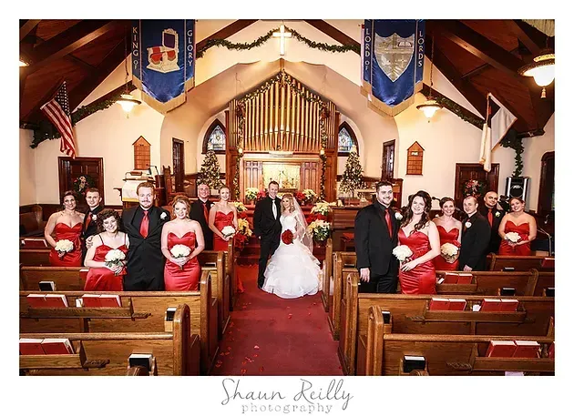 Wedding party posing in a church aisle, with bride and groom centered and bridesmaids in red dresses on both sides
