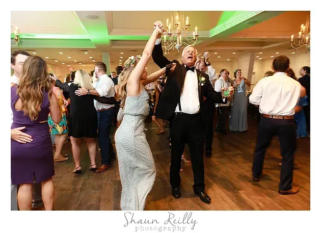 Guests dancing at a formal reception, with a woman in a silver dress and a man in a tuxedo in the center.