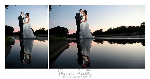 Bride and groom stand by a reflective pool at sunset, silhouetted against the evening sky.