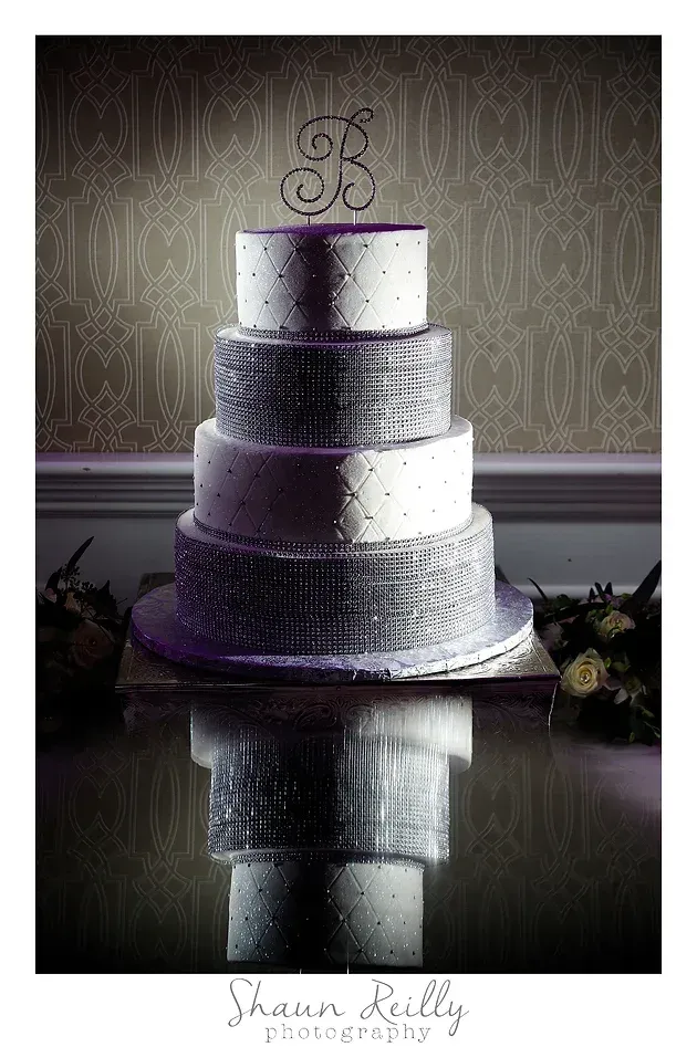 Four-tier white wedding cake with purple ribbon accents and monogram topper on a reflective table.