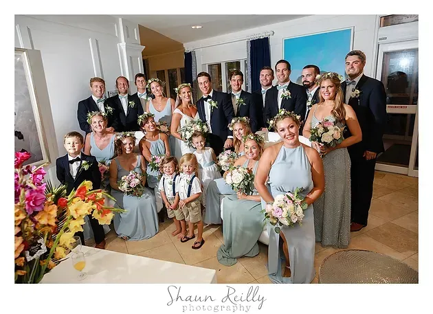 Formal wedding group posing indoors with bouquets and tuxedos, standing and seated in a decorated room