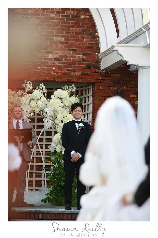 Bride in veil facing groom in black suit at wedding ceremony beside brick wall and floral arch