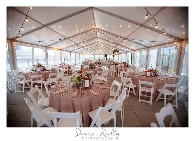 Elegant wedding reception tent with round tables, white chairs, blush tablecloths, and string lights