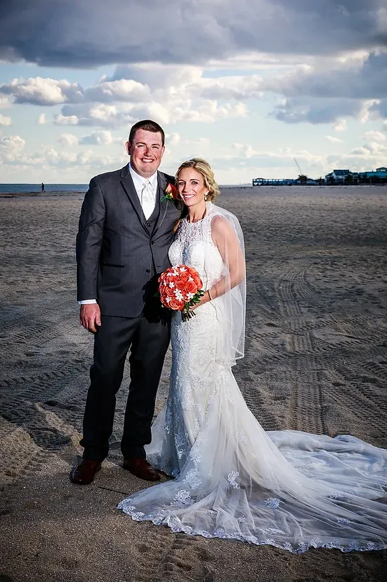 Newlywed couple posing on a beach, bride in white gown holding red bouquet under a cloudy sky