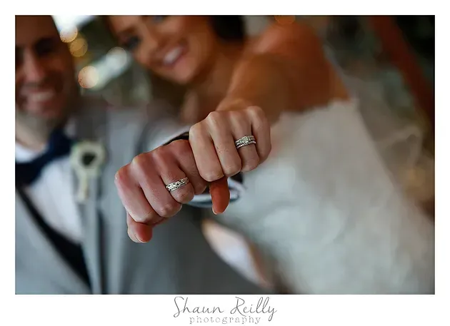 Close-up of a bride’s hand with wedding rings, held out in front of a softly blurred couple background