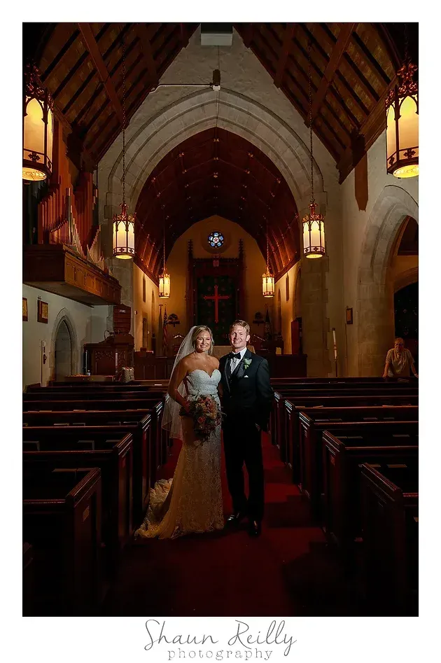 Bride and groom standing in a church aisle, with warm stained-glass light and a cross at the altar