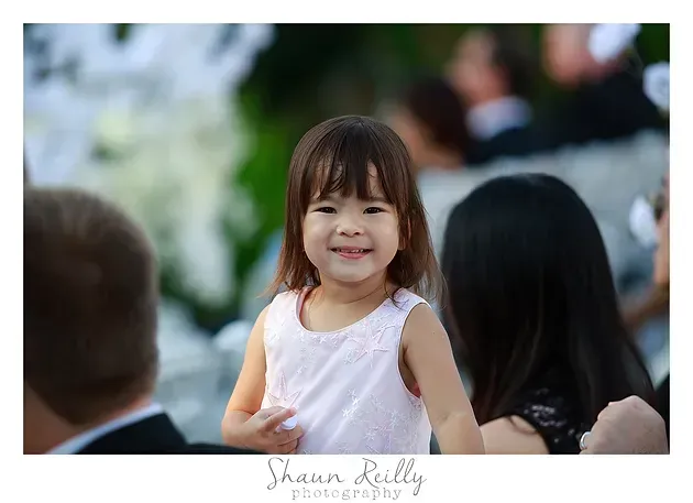 Smiling child in a white dress outdoors, with blurred people in the background