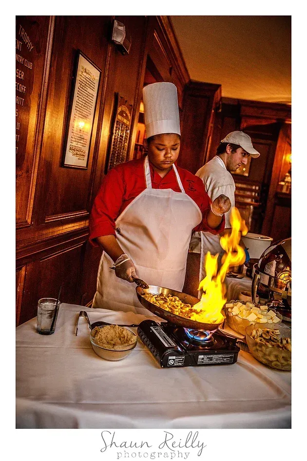 Chef in red jacket and tall hat flambés food at a restaurant table as another staff member watches.