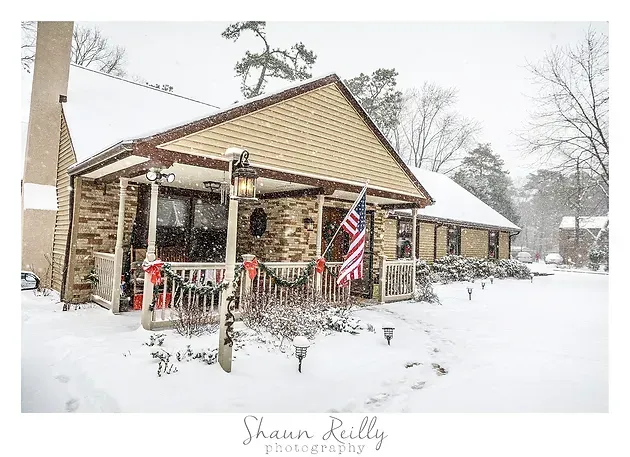 Snow-covered rustic cabin with a porch, American flags, and holiday decorations in a winter landscape