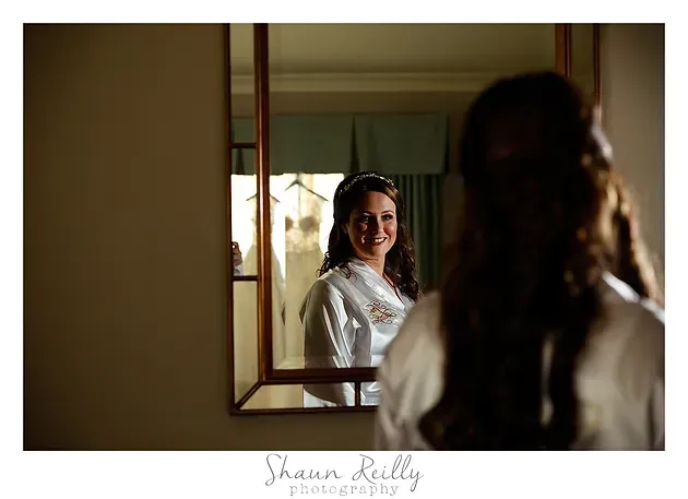 Woman smiling at her reflection in a mirror, wearing a white robe in a softly lit room