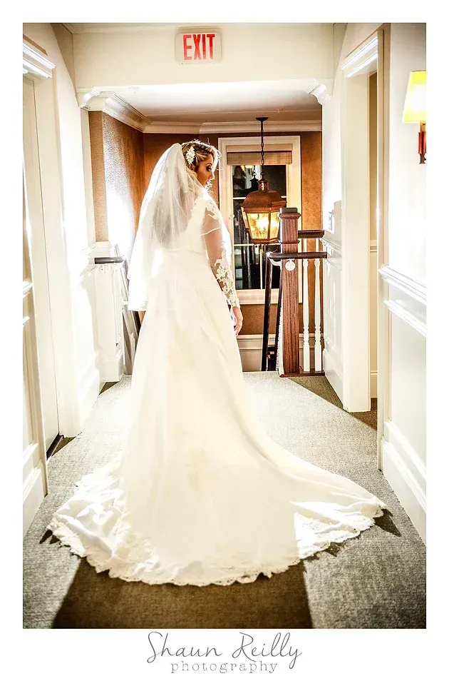Bride in a white gown and veil standing in a bright hallway with an exit sign behind her