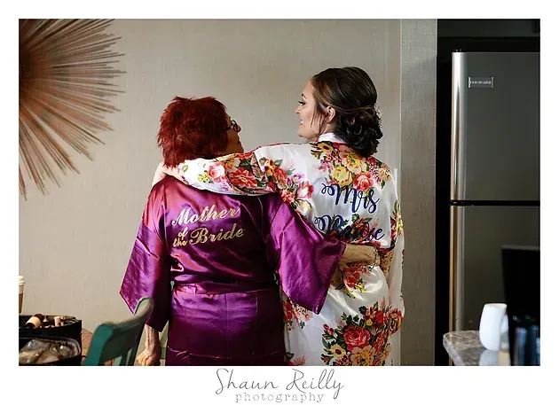 Two women in robes embrace in a kitchen beside a refrigerator.