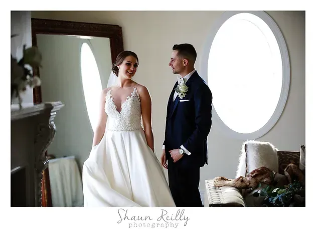Bride and groom standing together in a bright room, smiling at each other near an arched window.