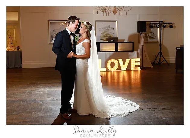 Bride and groom kiss in a ballroom, with glowing “LOVE” letters in the background.