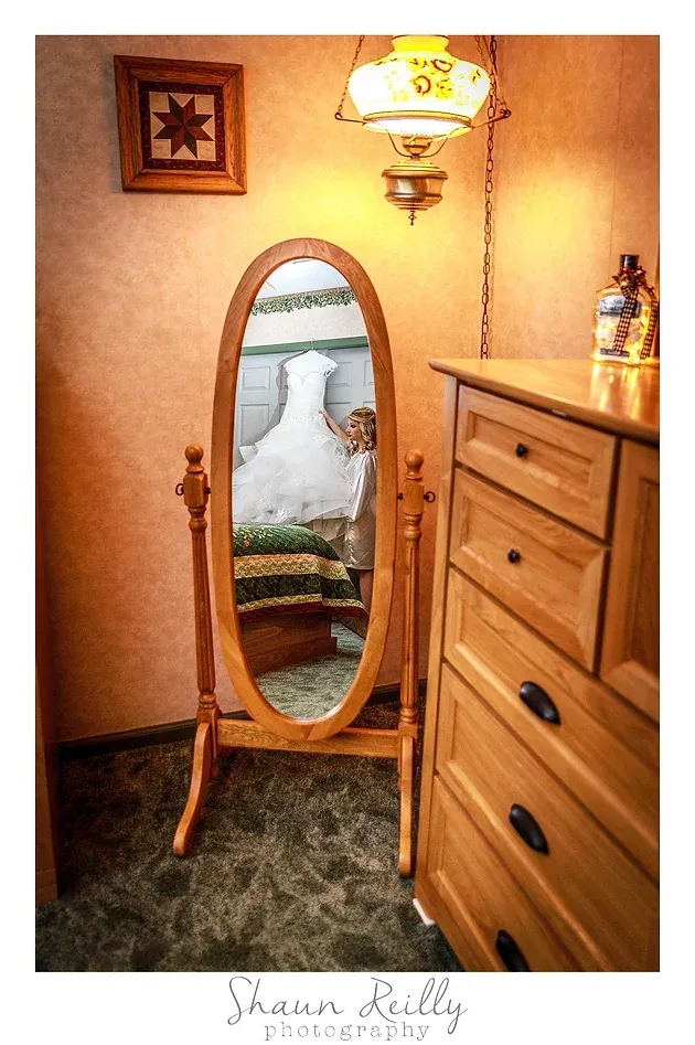 Oval mirror in a warmly lit bedroom reflecting a white bed and green blanket beside a wooden dresser.