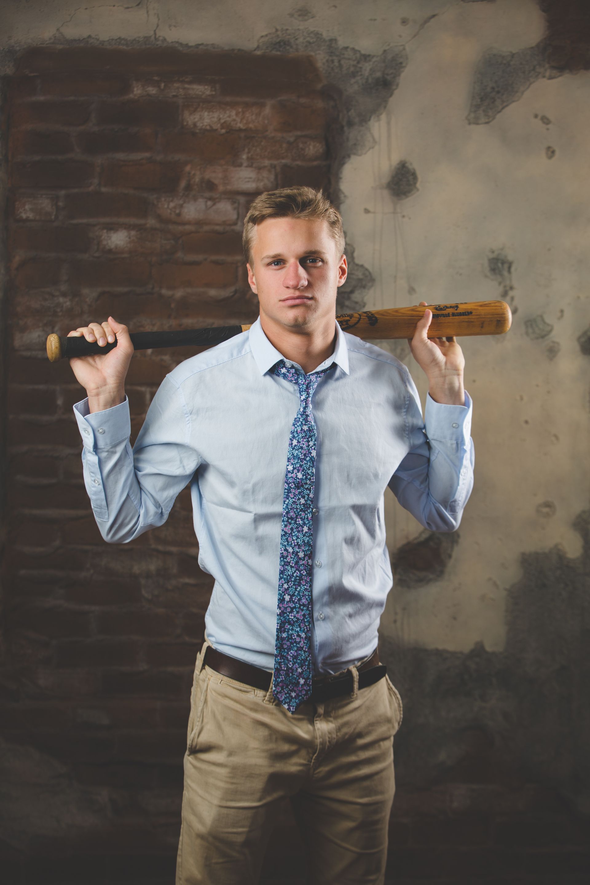 Man holding a wooden bat across his shoulders, wearing a blue shirt and tie against a brick wall.