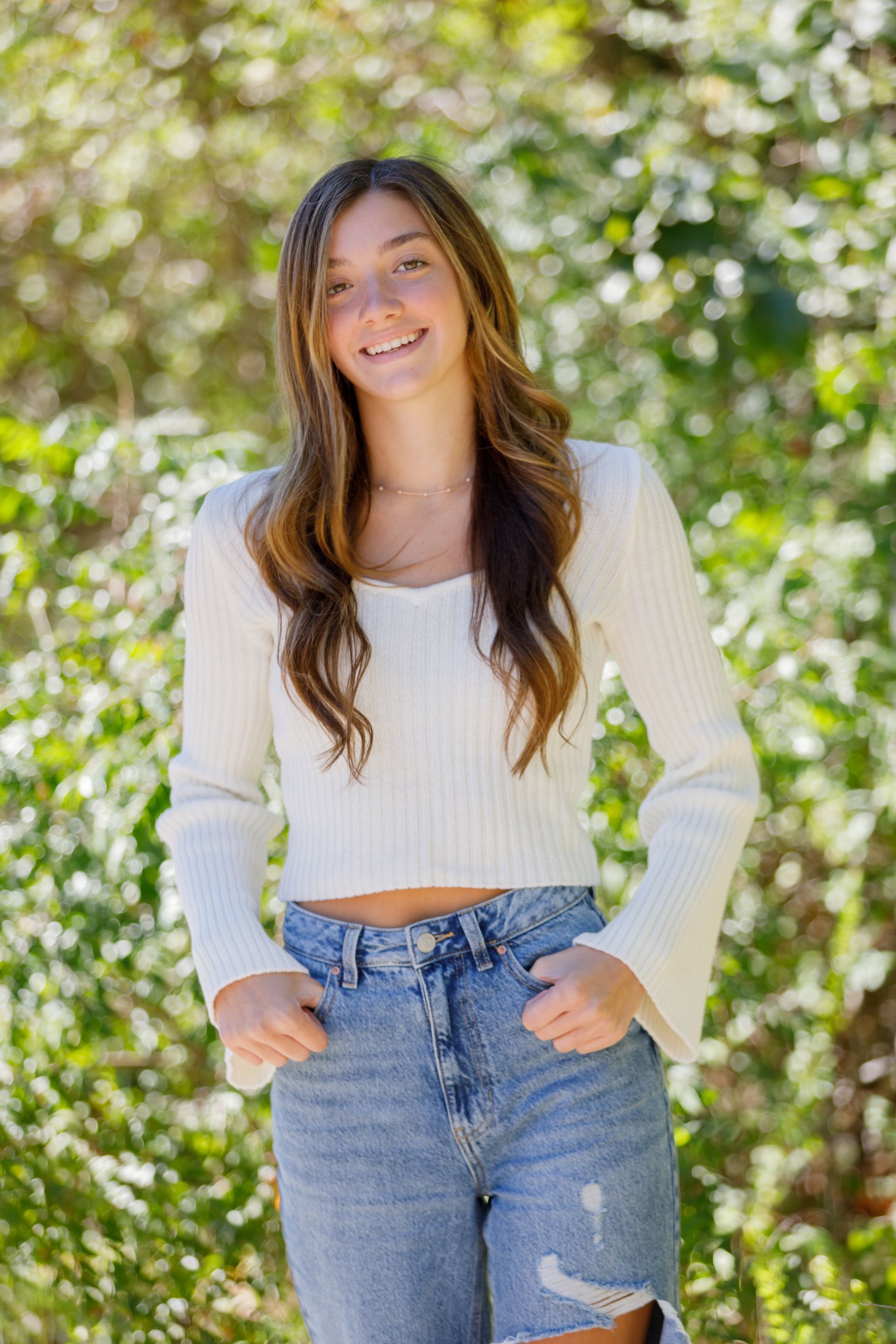 Smiling person in a white sweater and jeans standing outdoors in sunlight