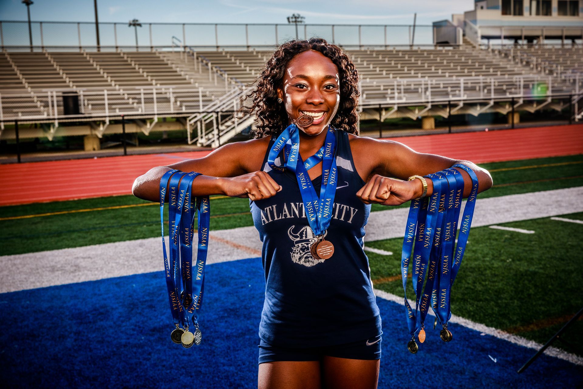 Athlete on a football field wearing medals, smiling and holding blue ribbons in front of stadium bleachers