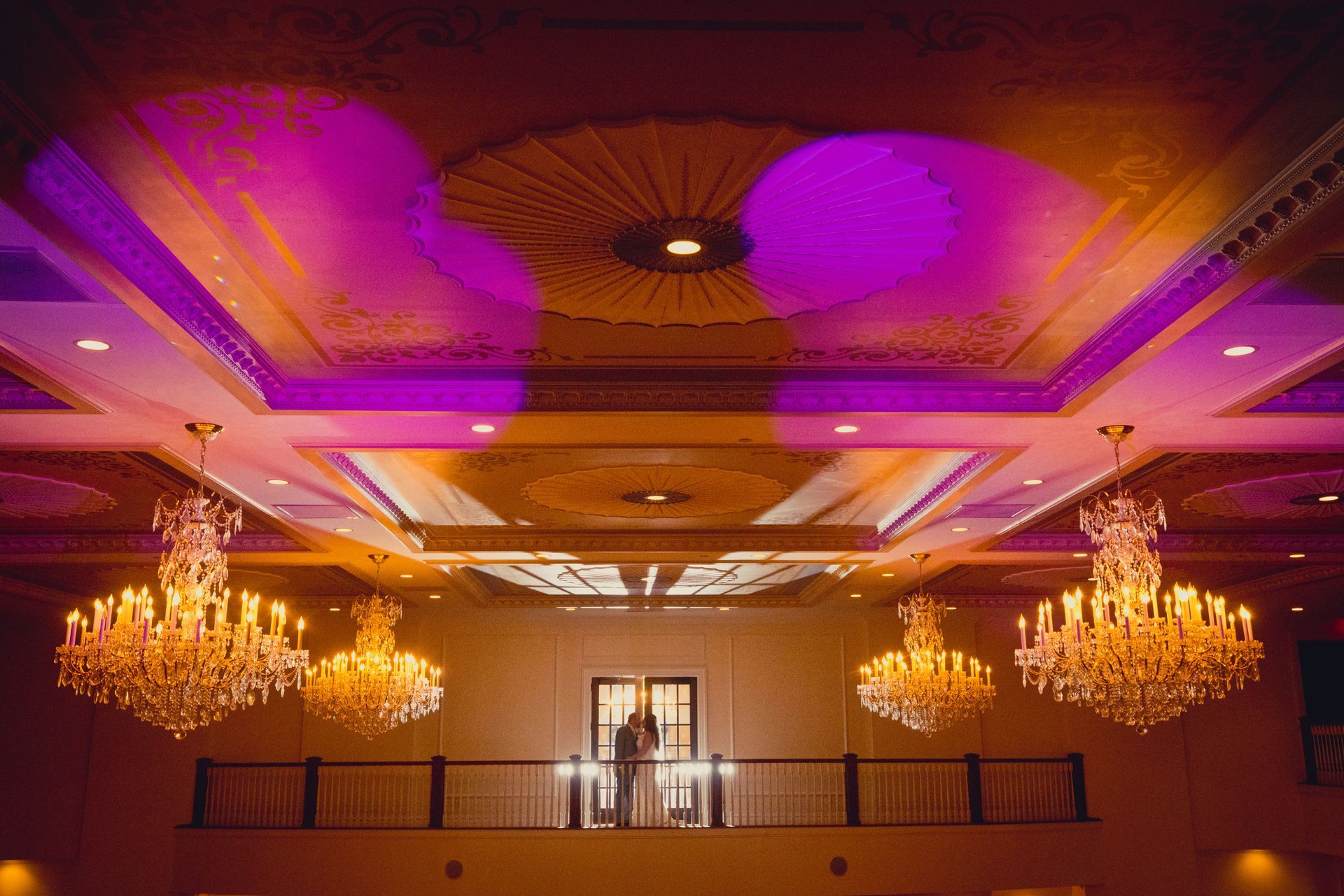 Grand ballroom ceiling lit with purple and gold chandeliers above a balcony railing