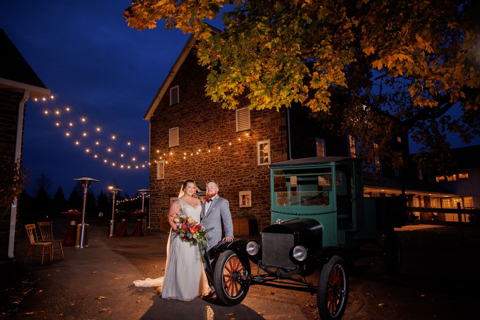 Bride and groom standing by a vintage car at a barn venue with string lights at dusk