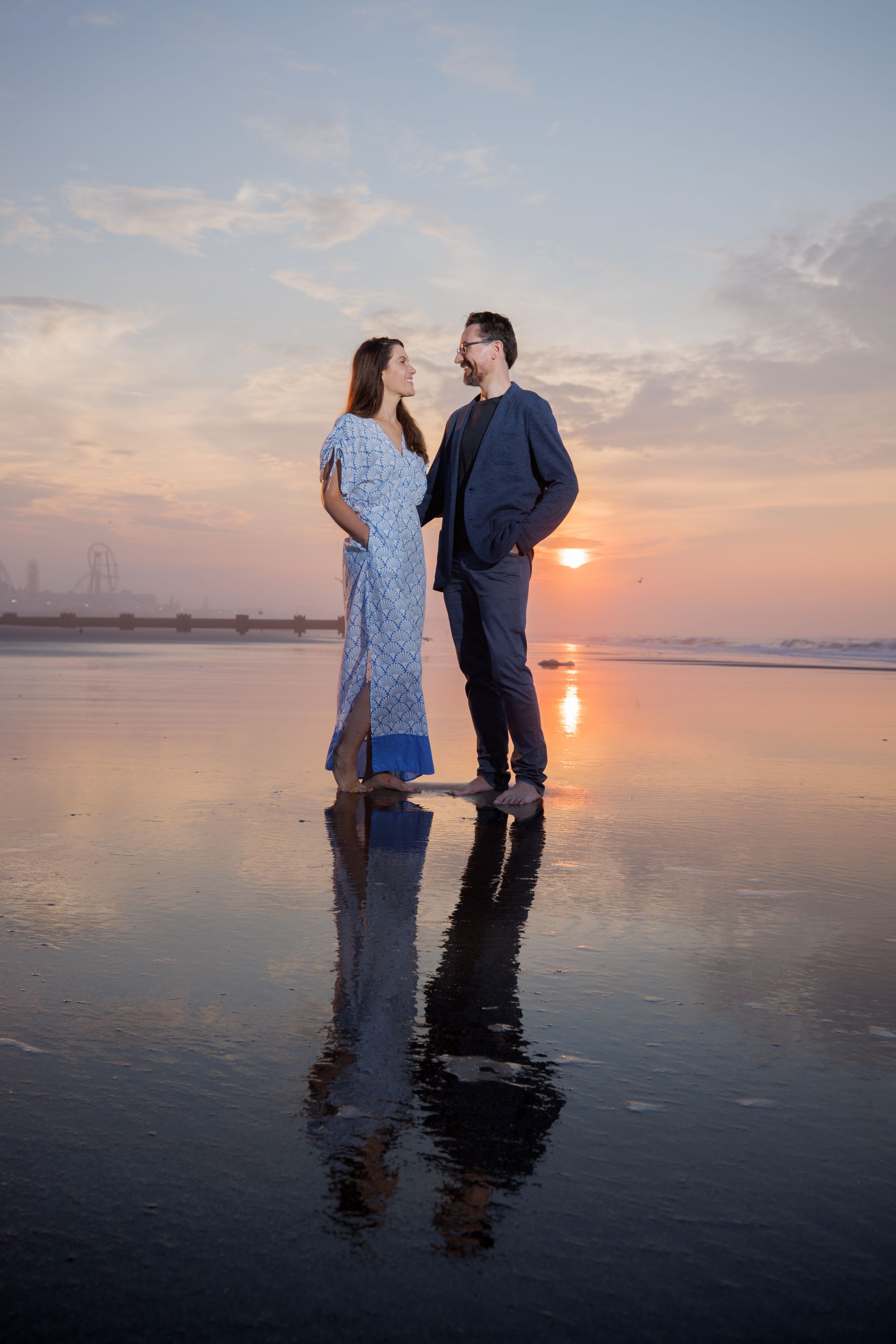 Two people standing on a reflective beach at sunset, facing each other near the water.