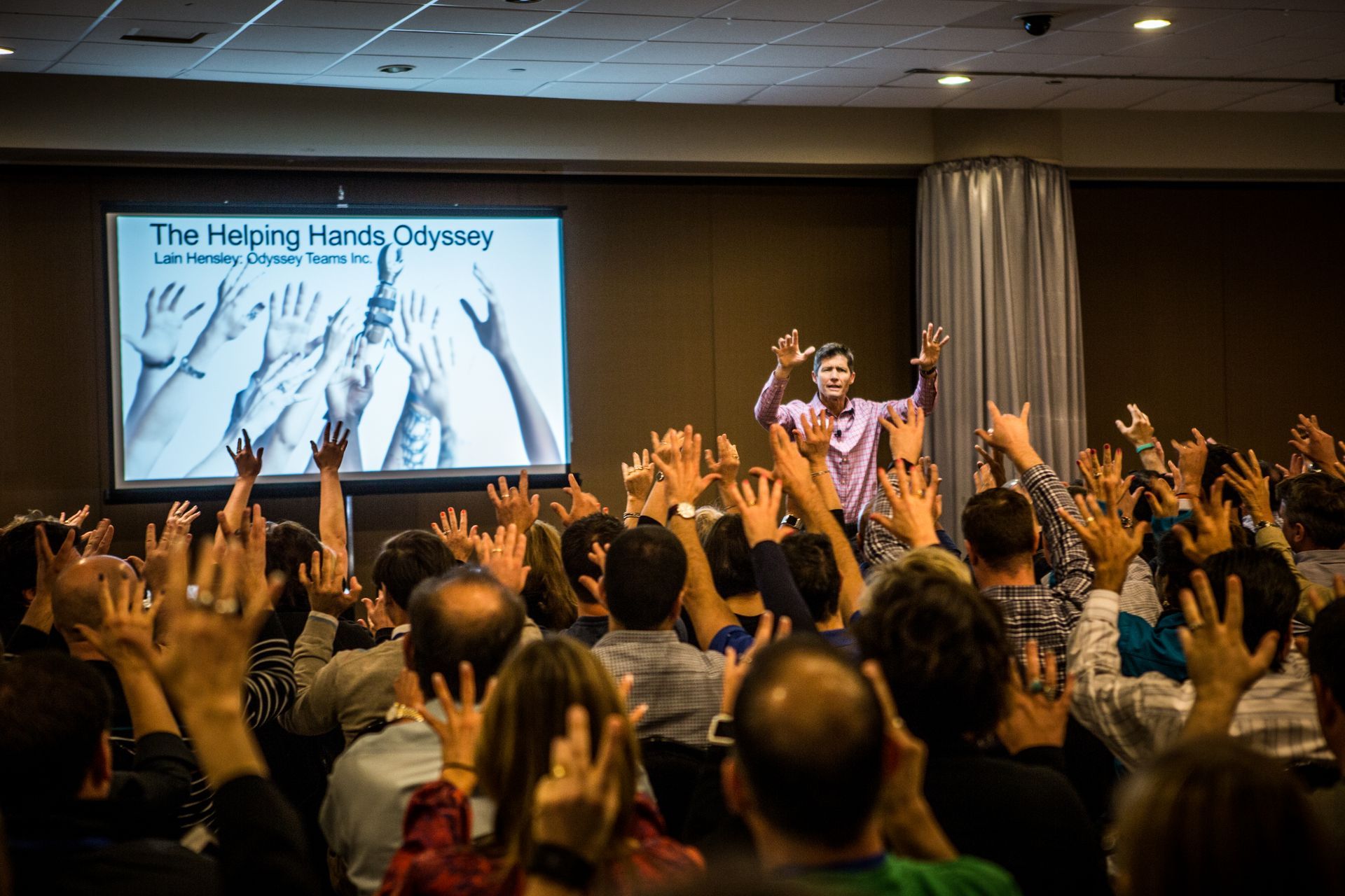 Audience raising hands at a conference presentation with a projected slide on stage