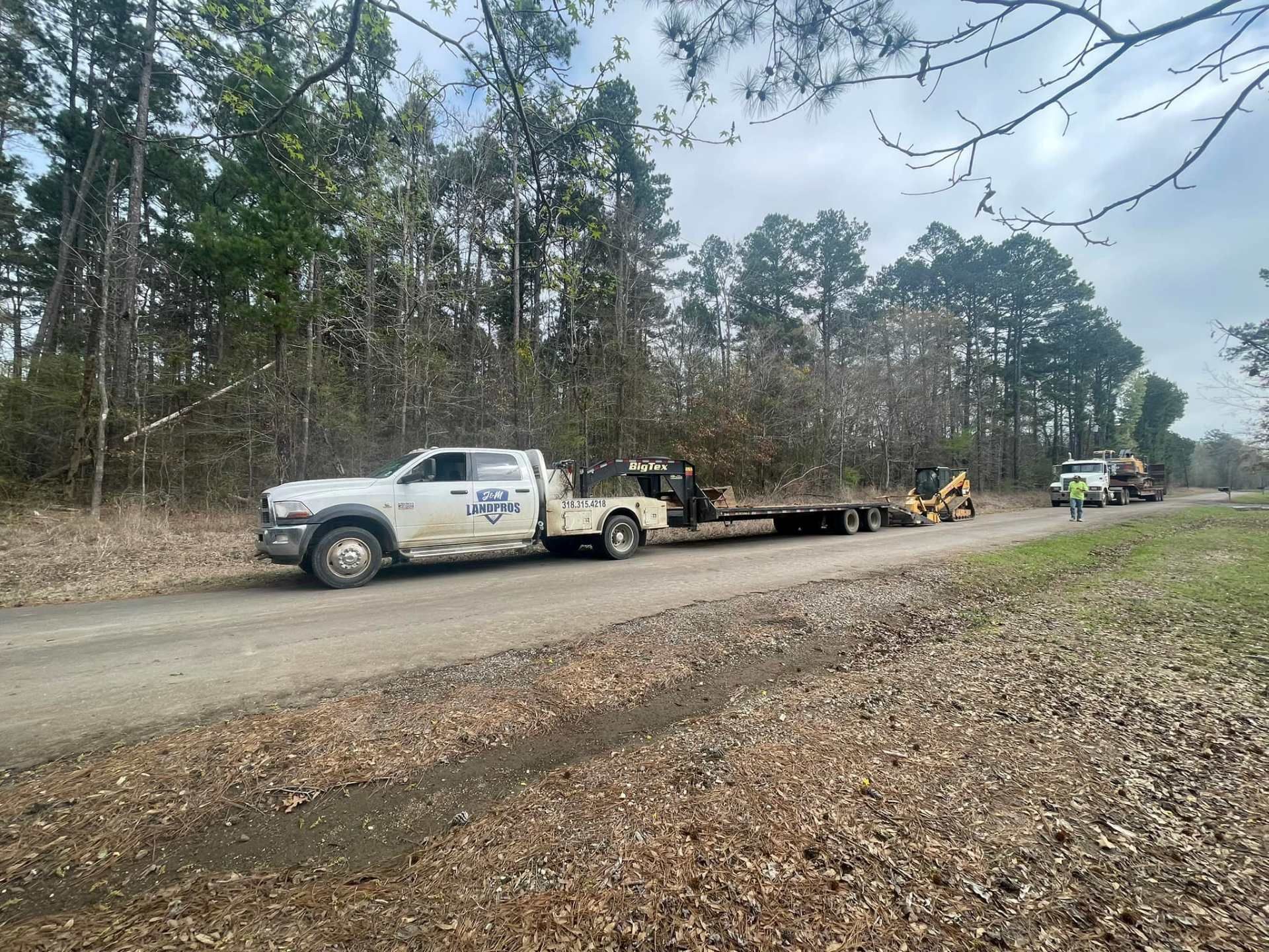 A white utility truck towing a flatbed trailer with a small piece of machinery on a dirt road near a wooded area.