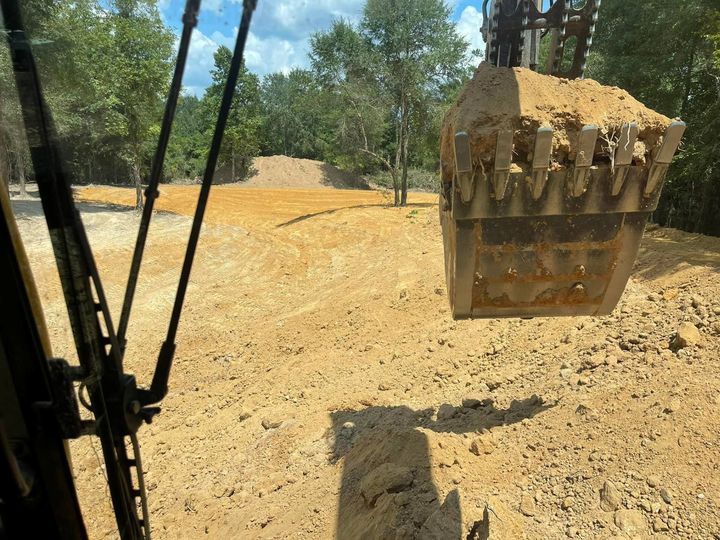The view from an excavator cab showing a large metal bucket filled with dirt, overlooking a cleared construction site.