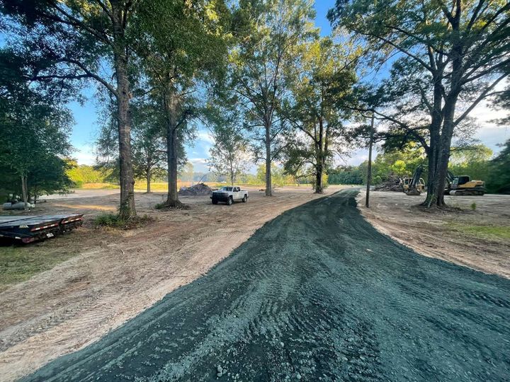 A gravel path curves through a wooded area toward an open field with a parked truck and construction equipment.